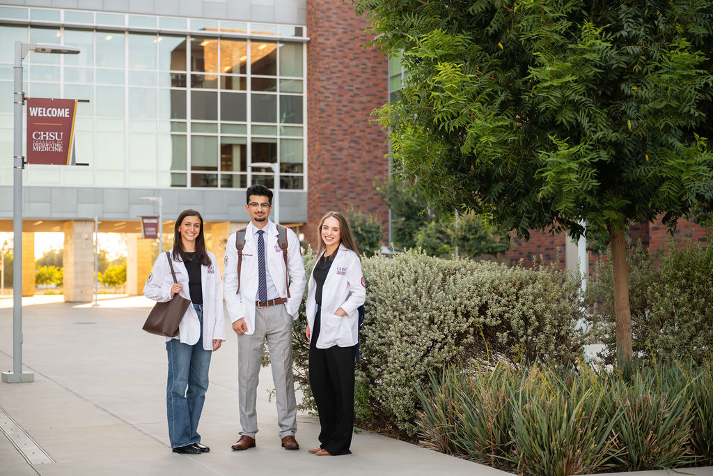 three students wearing white coats standing in courtyard at COM building.