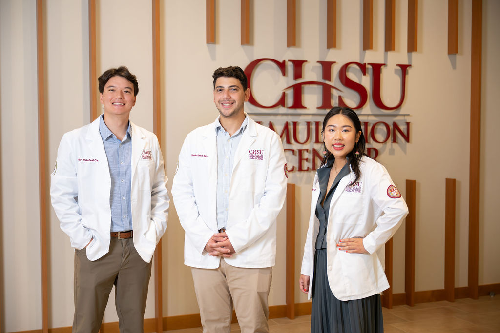 three students wearing white coats in front of CHSU Simulation Center sign