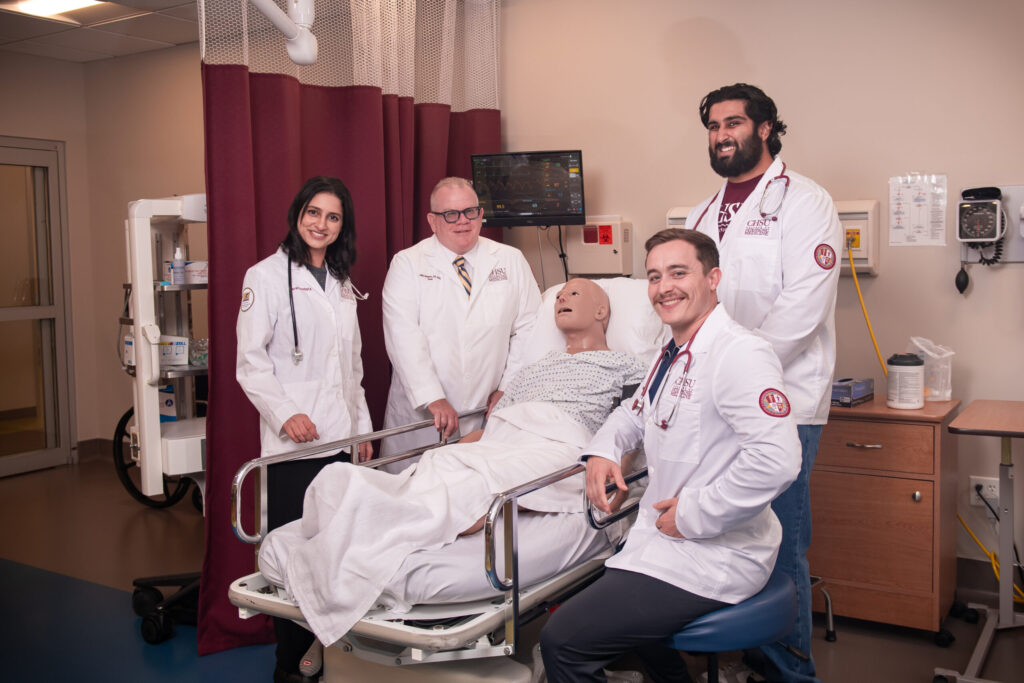 dean and students in white coats in hospital simulation room with mannikin in bed