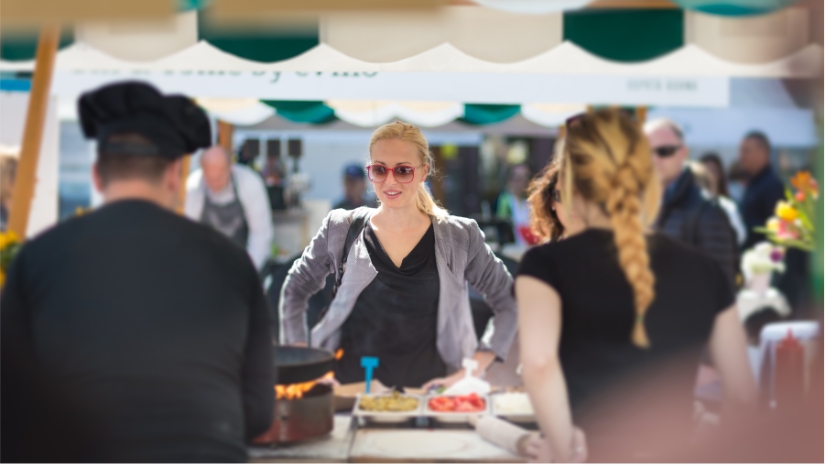 woman at food vendor tent looking at food with two people helping