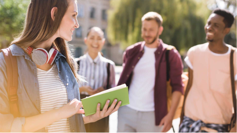 student holding book with headphones around neck smiling at 3 other students walking